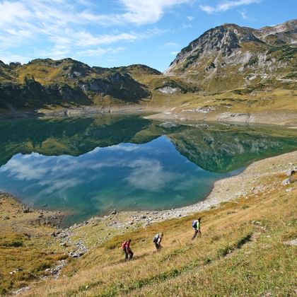 Drei Wanderer am Formarinesee auf dem Lechweg. Ein blauer Bergsee mit blauem Himmel und Hügellandschaft.