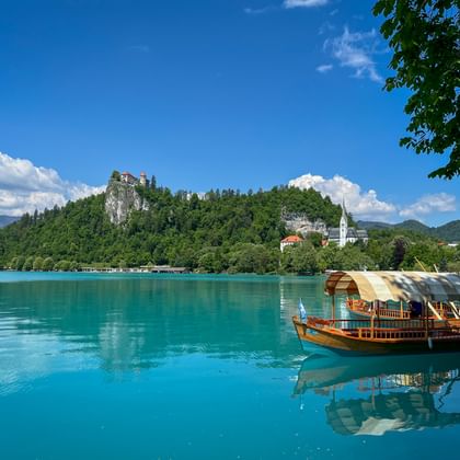 Traditionelles Holzboot auf dem türkisfarbenen Bleder See mit mittelalterlicher Burg auf bewaldetem Felsen und weißem Kirchturm am Ufer.
