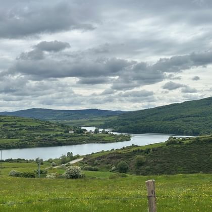 Fluss Ebro schlängelt sich durch üppige grüne Hügel bei Campoo de Polientes unter bewölktem Himmel. Wiese mit Wildblumen im Vordergrund.