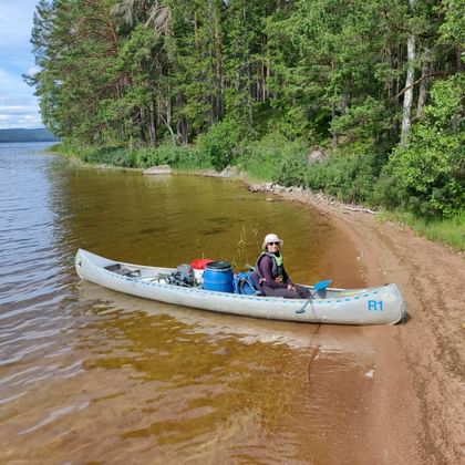 Frau im Kanu am schwedischen Seeufer Frau sitzt in weißem Kanu am sandigen Ufer eines schwedischen Sees, umgeben von dichtem Wald. Klares Wasser und blauer Himmel sichtbar.