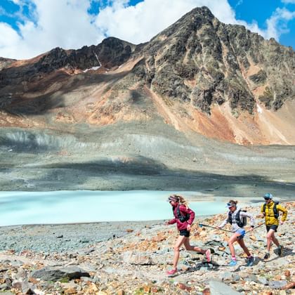Drei Trailrunner joggen uber einen schmalen Natursteinweg neben einem Bergsee über die via Grischuna. Im Hintergrund der massive Berg unter leicht bewölktem blauen Himmel.