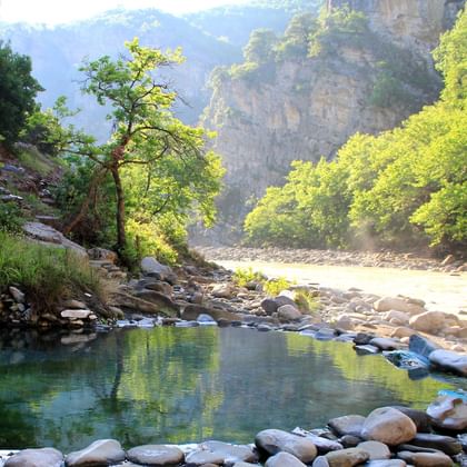 Klarer Bergpool umgeben von glatten Steinen in albanischer Schlucht. Steile Klippen mit üppiger grüner Vegetation erheben sich beiderseits unter nebligen Bergen.