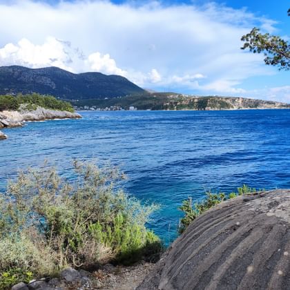 Tiefblaue Bucht in Albanien mit felsiger Küste, Bergen im Hintergrund und mediterraner Vegetation im Vordergrund unter teilweise bewölktem Himmel.