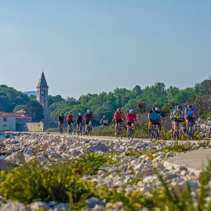 Radfahrergruppe auf Küstenradweg entlang der Kvarner Bucht mit Steinmole, historischem Glockenturm und Gebäuden am gegenüberliegenden Ufer.