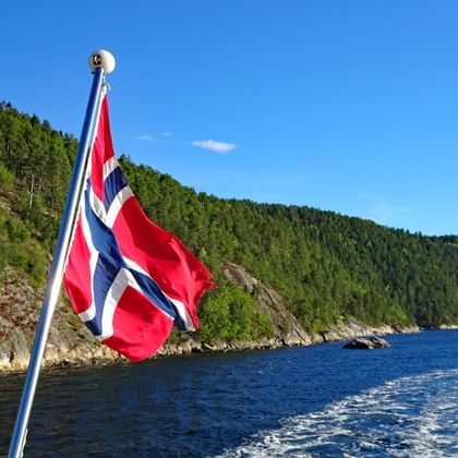 Drapeau norvégien flottant depuis un bateau sur l'Oslofjord avec collines boisées et côte rocheuse. Eau bleue et ciel clair en arrière-plan.