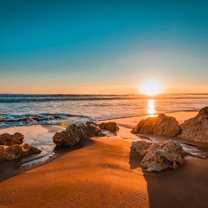 Goldener Sonnenuntergang über portugiesischem Strand mit großen Felsen im Sand. Wellen spülen das Ufer während die Sonne warmes Licht aufs Wasser wirft.