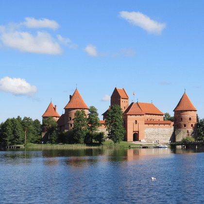 Rote Backsteinburg Trakai mit mehreren Türmen spiegelt sich in ruhigem Seewasser, umgeben von grünen Bäumen unter blauem Himmel.