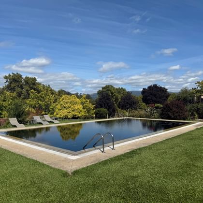 Großer rechteckiger Pool mit Steinumrandung auf gepflegtem Rasen im Hotel Convento dos Capuchos, umgeben von Bäumen unter blauem Himmel.