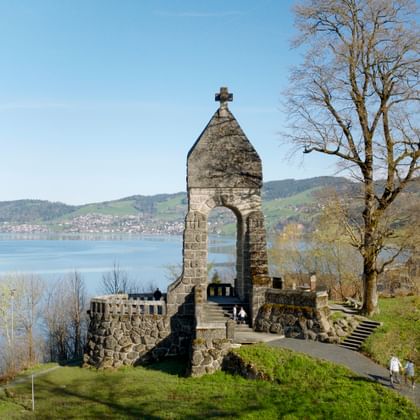 Stone tower with cross at Morgarten Monument overlooking Lake Aegeri. The memorial features a chapel tower and battlements on a green hillside.