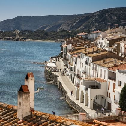 Weiße Häuser mit Terrakottadächern säumen die Uferpromenade eines mediterranen Küstendorfs, mit blauem Meer und grünen Hügeln im Hintergrund.