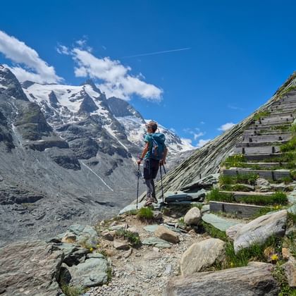 Wanderin mit Rucksack und Stöcken auf Bergpfad neben Holzstufen, vor schneebedeckten Gletschergipfeln unter blauem Himmel mit Wolken.