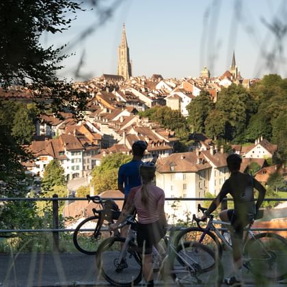Three cyclists with gravel bikes stand at a viewpoint overlooking Bern's old town with church spires and the Aare river winding through.