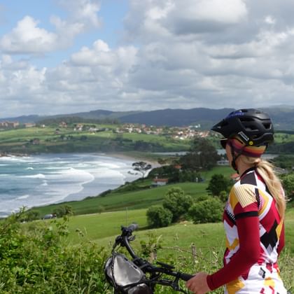 Radfahrerin mit Helm und buntem Trikot mit Blick auf den Oyambre Strand und Küstenlandschaft mit grünen Hügeln und bewölktem Himmel.