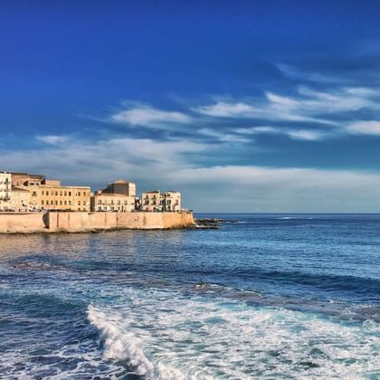 Vue côtière de Syracuse, Sicile montrant des bâtiments historiques en pierre le long du front de mer avec la mer Méditerranée bleue.