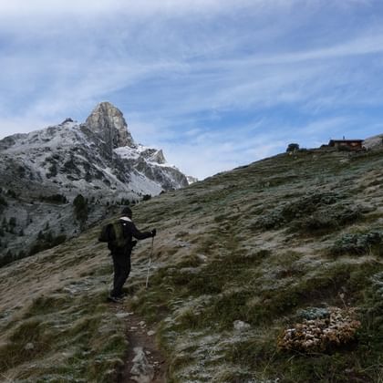 Wanderer mit Rucksack und Stöcken auf Alpenpfad mit verschneitem Meidhorn-Gipfel und Berghütte. Frost bedeckt das Gras entlang des Alpenpässewegs.