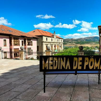 Schwarze Bank mit 'MEDINA DE POMAR' in goldenen Buchstaben auf einem Platz, mit bunten historischen Gebäuden und Bergen unter blauem Himmel im Hintergrund.