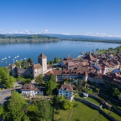 Luftaufnahme der mittelalterlichen Stadt Murten mit Schlossturm am Murtensee. Historische Gebäude mit roten Dächern säumen das Seeufer.