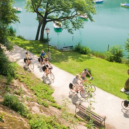 Gruppe von Radfahrern auf Seeweg am Mattsee mit türkisfarbenem Wasser, Booten und grüner Rasenfläche umgeben von Bäumen.