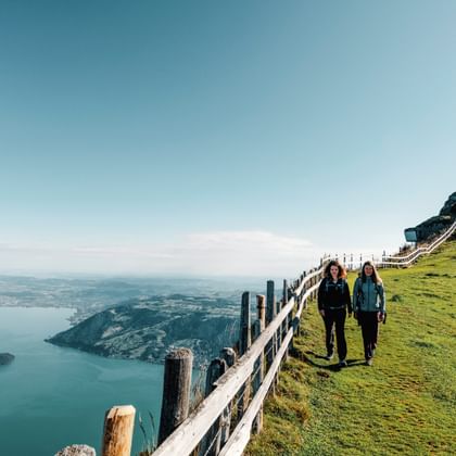 Two hikers walking on Mount Rigi's grassy summit with wooden fence, transmission tower, and panoramic view of Lake Lucerne below.