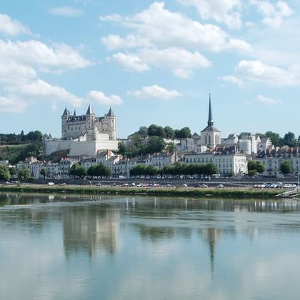 Château blanc et ville se reflétant dans la Loire calme sous un ciel bleu avec des nuages blancs. Les bâtiments historiques bordent la rive.