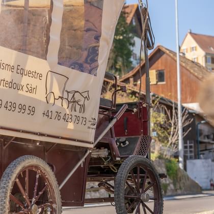 Traditioneller Planwagen mit weißer Plane und Schild 'Tourisme Equestre Courtedoux Sàrl', geparkt in einer Dorfstraße im Jura Ajoie.