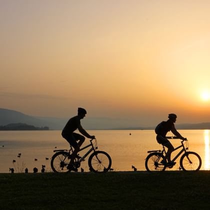Deux cyclistes en silhouette contre le ciel doré du coucher du soleil près d'un lac calme dans la région de Bienne Seeland, avec des collines en arrière-plan.
