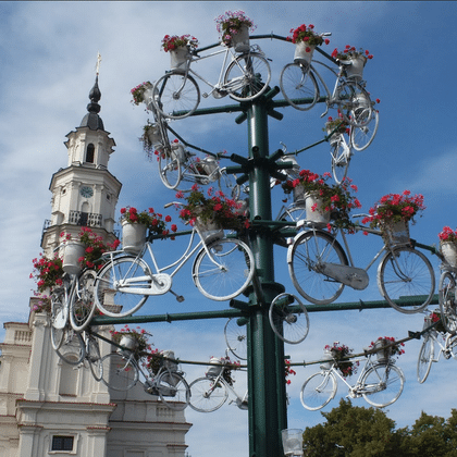 Hoher Mast dekoriert mit weißen Fahrrädern und bunten Blumenkörben vor blauem Himmel. Historischer Kirchturm mit Kuppel im Hintergrund.