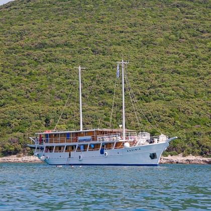 Weiße Segelyacht MS Kazimir mit zwei Masten vor Anker in ruhigen blauen Gewässern nahe einer bewaldeten Küste.