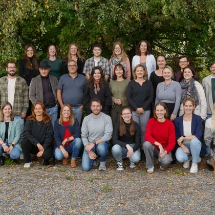 Group of approximately 25 people arranged in three rows for a team photo outdoors, with green foliage background and a dog in the front row.