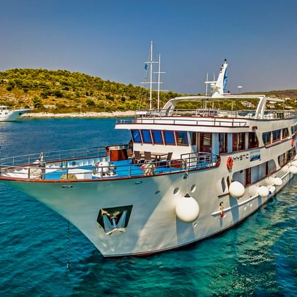 Weißes Kreuzfahrtschiff MY Mystic vor Anker in kristallklarem türkisem Wasser nahe einer bewaldeten Küste mit anderen Schiffen im Hintergrund.