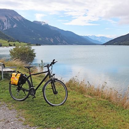 Vélo noir avec sacoches jaunes garé sur l'herbe au bord du lac de Reschen, entouré de montagnes et prairies vertes sous un ciel nuageux.