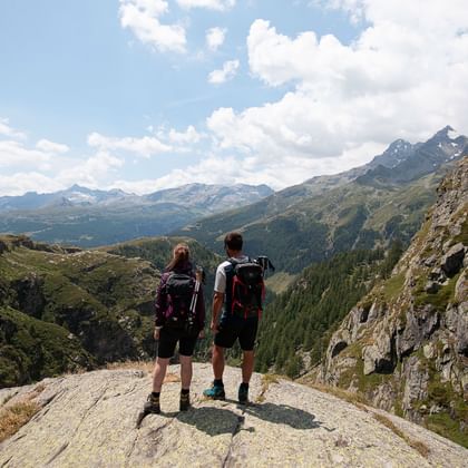 Ein Wanderer-Pärchen steht auf einem Steinvorsprung und schaut sich die umliegenden Berge an.