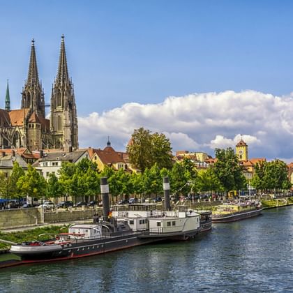 Regensburger Dom mit gotischen Zwillingstürmen über der Donau. Historische Boote am Ufer mit bunten Gebäuden und Bäumen.