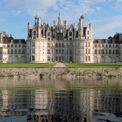 Le château de Chambord avec ses tours et cheminées distinctives se reflète dans la Loire, entouré de pelouses vertes sous un ciel bleu.