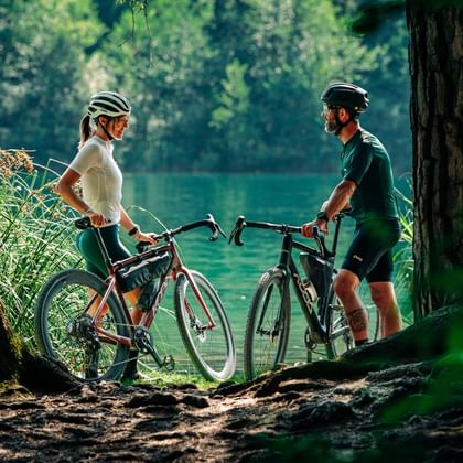 Zwei Radfahrer mit Helmen und Gravelbikes pausieren an einem türkisen See in Graubünden, umrahmt von Bäumen und üppiger Vegetation.