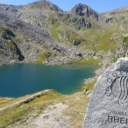 Marqueur des sources du Rhin au lac alpin Marqueur en pierre indiquant 'Quelle Rhein' près d'un lac alpin turquoise entouré de montagnes rocheuses. Une tente rouge est visible sur la rive.