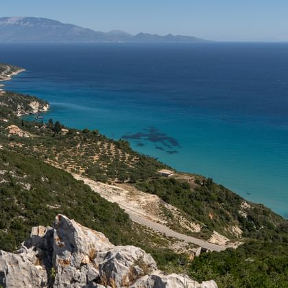 Luftaufnahme der Zakynthos-Küste mit türkisfarbenem Mittelmeer, grünen Hügeln, weißen Klippen und fernen Bergen unter blauem Himmel.