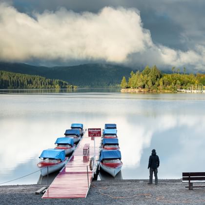 Person standing at Schluchsee lake shore beside a wooden dock with moored rowing boats. Forested mountains and dramatic clouds reflect in calm water.