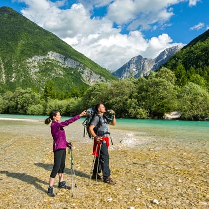 Wanderpaar mit Fernglas am steinigen Ufer der türkisen Soca, mit den Bergen im Hintergrund