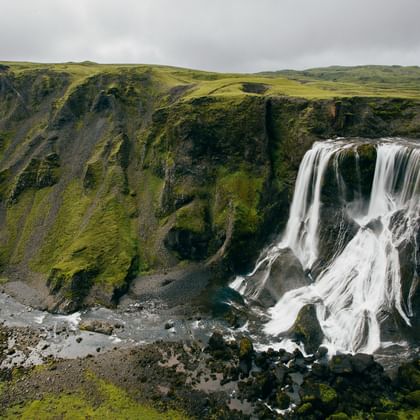 Das ganze Bild ist von eine grünen Klippe gefüllt von der auf der rechten Seite ein grosser Wasserfall herunter fliesst. Der Himmel ist grau und bewölkt.