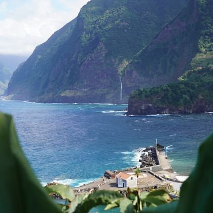 Küstensiedlung mit Pier und weißer Kapelle auf Madeira, eingerahmt von grünen Blättern. Steile Klippen und Wasserfall zum türkisen Ozean.