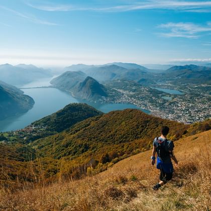 Wanderer mit Rucksack auf goldenem Grasland am Monte Boglia mit Panoramablick auf den Luganer See und die umliegenden Berge im Tessin.