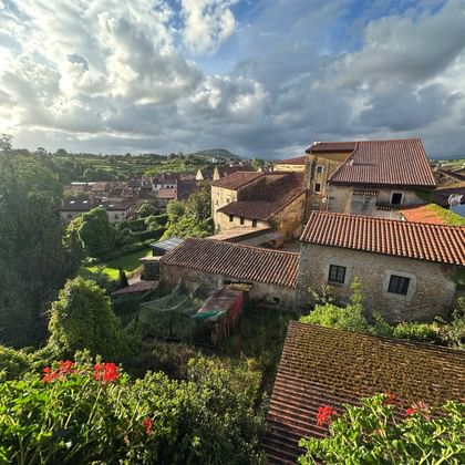 Steinhäuser mit Terrakottadächern in Santillana del Mar unter dramatischem Wolkenhimmel. Rote Blumen im Vordergrund, grüne Hügel in der Ferne.