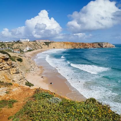 Plage pittoresque d'Aljezur avec falaises de grès doré, vagues turquoise, rivage sablonneux et baigneurs sous ciel bleu.