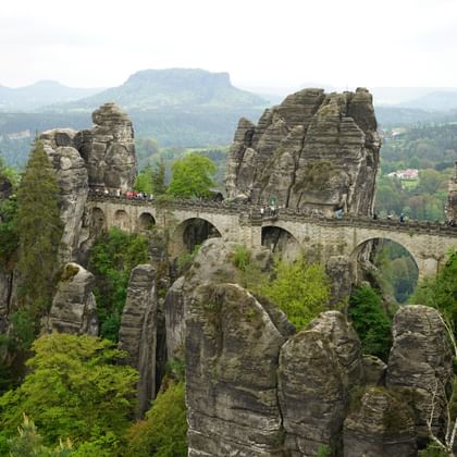 Historische Steinbrücke verbindet hohe Sandsteinfelsen, umgeben von grünem Wald mit fernen Bergketten im Hintergrund.