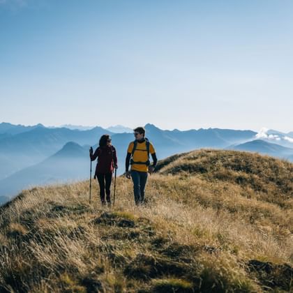 Zwei Wanderer mit Rucksäcken auf einem grasbewachsenen Bergrücken. Gestaffelte Bergketten und Wolken in der Ferne unter blauem Himmel.