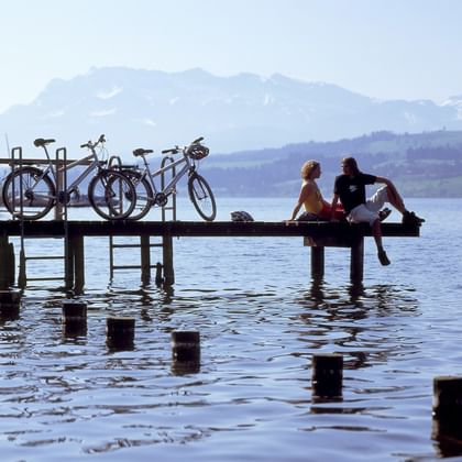 Zwei Radfahrer sitzen auf Holzsteg mit Fahrrädern und blicken auf Schweizer See mit Bergen im Hintergrund. Holzpfosten im Wasser sichtbar.