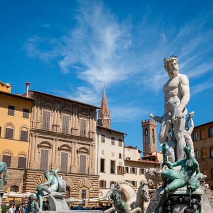 Fontaine de Neptune à Florence avec statue en marbre blanc et figures en bronze, entourée de bâtiments historiques et clochers sous ciel bleu.