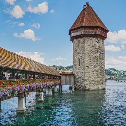 Le Pont de la Chapelle à Lucerne avec sa tour d'eau en pierre emblématique, décoré de fleurs colorées, enjambant l'eau bleue sous un ciel nuageux.