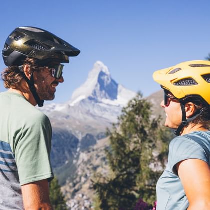 Two cyclists stand in front of the Matterhorn and look at each other.